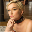 Load image into Gallery viewer, A close-up of a blonde woman wearing the Leather Buckling Collar with Scalloped Edges is shown. The collar is made of a wide piece of black leather with a smaller notched strip wrapped around it. It has red scalloped trim and metal hardware.
