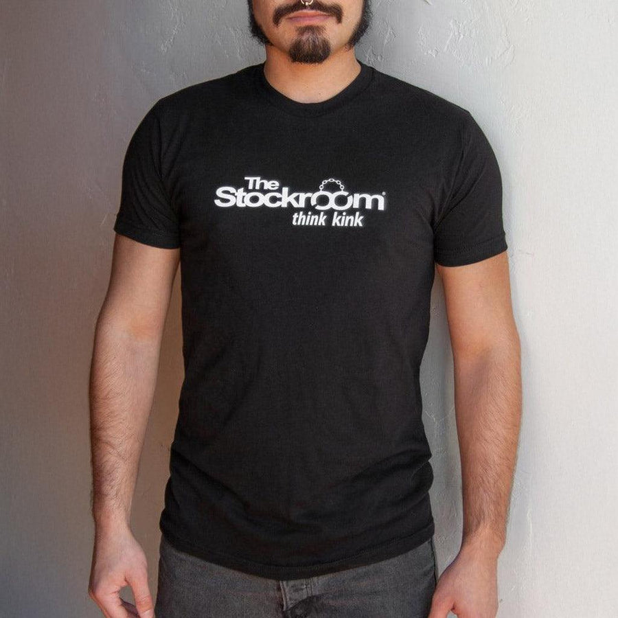 A man is shown wearing a Men's Stockroom Think Kink T-Shirt. The T-shirt is black and has the Stockroom logo printed in white on it, with the slogan “Think Kink” underneath.