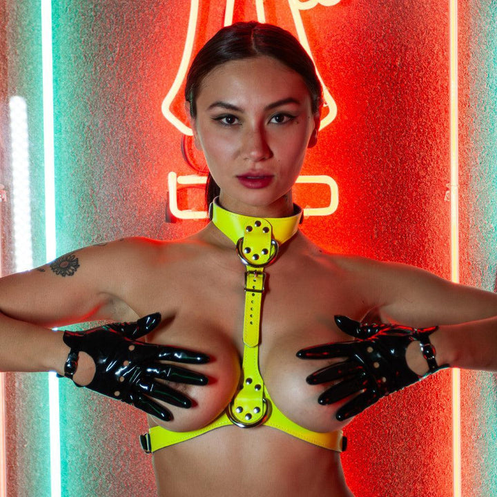 A nude woman with her hair pulled back wears the yellow Neon Angel Bust Harness and black latex biker gloves. She is standing in front of a glowing neon sign and covers her breasts with her hands.