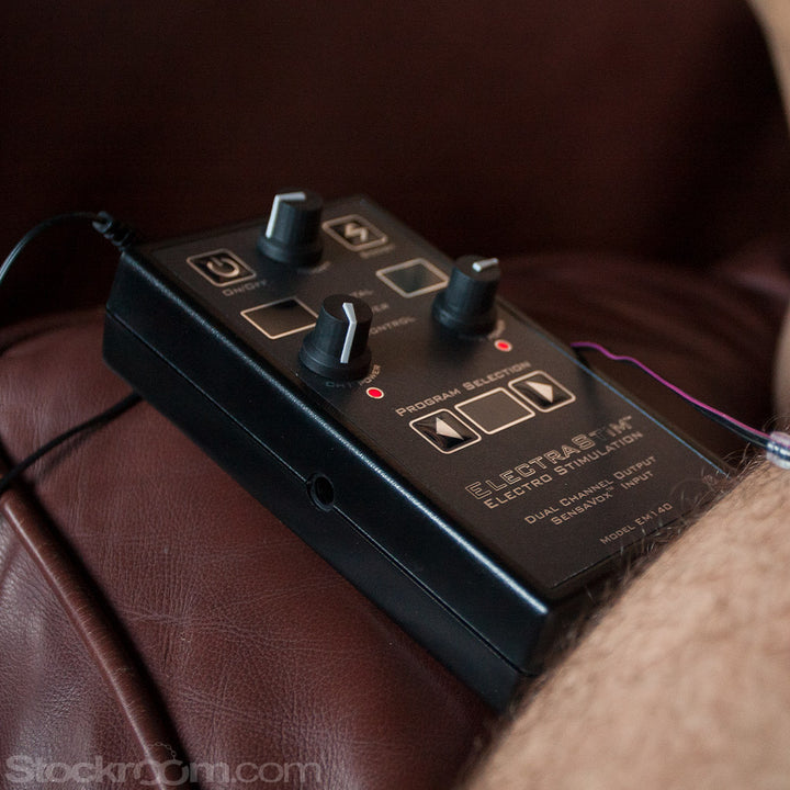 A close-up of an unidentifiable part of a man’s body is shown resting on a brown leather couch. On his leg rests a red silicone pinwheel. The pinwheel is plugged into the Electrastim Sensavox, a grey box with three dials on it and multiple buttons. 