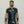Load image into Gallery viewer, A brunette man with facial hair poses in front of a white wall. He wears the Uniform Shirt from Syren Latex in black, matching pants, and arm gauntlets. The shirt has metal snaps down the front, pockets on the chest, and a collar.

